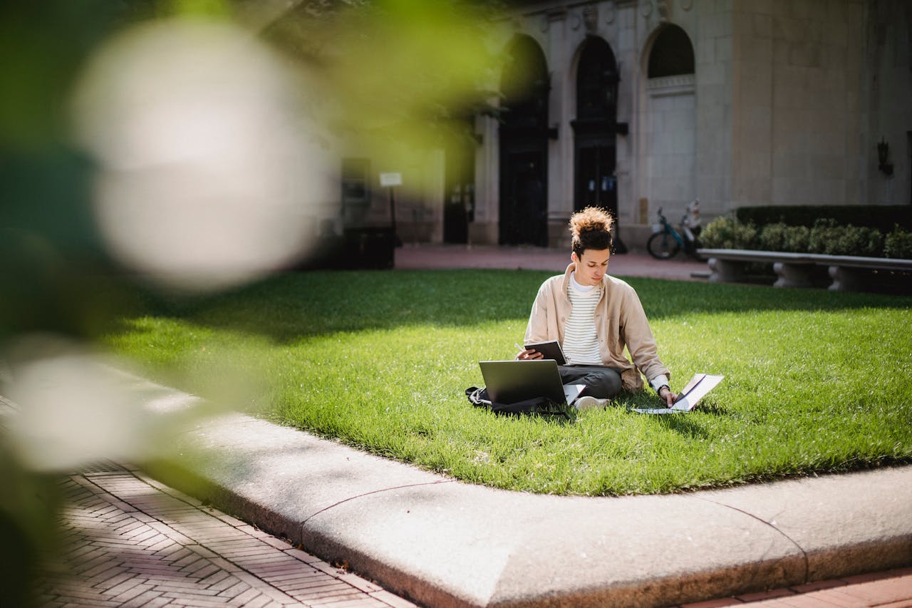 A young man studies with a laptop and books on a sunny university campus lawn.
